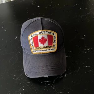 Svart keps från Dsquared2 med patch - Svart keps från Dsquared2 med stor broderad patch framtill med kanadensisk lönnlöv och text. Justerbar rem bak och broderad text ovanför öppningen. Klassisk böjd skärm och cool streetstil. 