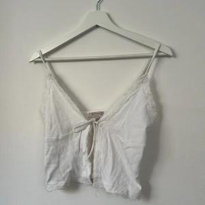 Vit ribbad linnetopp från Pull&Bear - Supersöt vit ribbad linnetopp från Pull&Bear med smala axelband, spetsdetaljer längs kanterna och knyt framtill. Croppad modell som ger en fräsch och somrig vibe. Perfekt att matcha med jeans eller kjol för en chill look.