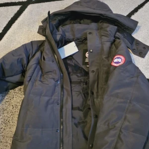 Svart dunjacka från Canada Goose - Säljer en svart dunjacka från Canada Goose med huva och avtagbar pälskant. Jackan har klassisk patch på ärmen, dragkedja och knappar framtill. Perfekt för kalla dagar och riktigt snygg streetstil.