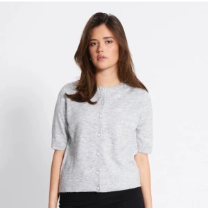Ljusgrå stickad kofta med pärlknappar - Superfin ljusgrå stickad kofta med korta ärmar och pärlknappar framtill. Mjuk och fluffig känsla, perfekt för att addera lite cozy vibes till din outfit. Rund halsringning och ribbade kanter ger en clean look.
