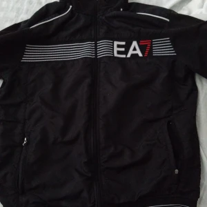 Svart vindjacka EA7 Emporio Armani - Svart vindjacka från EA7 Emporio Armani med vita och röda detaljer samt logga på bröstet och ärmen. Jackan har dragkedja framtill, ståkrage och sidofickor med dragkedja. Snygga ränder över bröstet ger en sportig vibe.