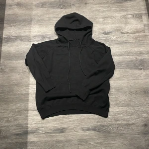 Svart merinoull hoodie med huva och snörning - En svart merinoull hoodie med huva och snörning framtill. Tröjan har lång ärm och ribbade muddar vid ärmslut och nederkant. Enkel och stilren design som passar till det mesta. Materialet känns mjukt och bekvämt.