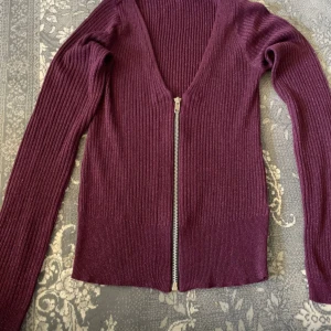 Mörklila ribbad kofta med dragkedja - Säljer en mörklila ribbad kofta med v-ringning och hel dragkedja framtill. Tröjan är stickad och har långa ärmar, perfekt för lager på lager. Snygg och enkel att matcha till olika outfits.