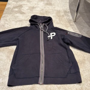 Svart hoodie från Pelle P med dragkedja - Mörk hoodie från Pelle P med hel dragkedja framtill, broderad logga på bröstet och praktisk ficka med dragkedja på ärmen. Tröjan har huva med snörning och stora fickor framtill. Perfekt för kyliga dagar och snygg till jeans.