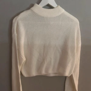 Vit stickad croppad tröja - Säljer en vit stickad tröja med croppad passform och lång ärm. Tröjan har en högre ribbad halsringning och är perfekt för lager-på-lager-stil under kyliga dagar.