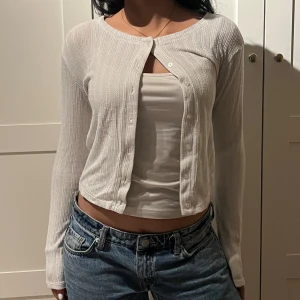 Vit ribbad croppad topp med knappar - Säljer en vit croppad topp med ribbad struktur och långa ärmar. Toppen har knappar framtill och en rund halsringning. Perfekt att styla med jeans för en chill look.