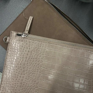 Beige kuvertväska i skinnimitation - 2 stycken kuvertväskor från carin wester, första är en beige kuvertväska med krokodilpräglad yta och silvrig dragkedja.  Den andra väskan  är i en mockaimitation och detaljer i samma färg. Perfekt för att bära det viktigaste med snygg touch.