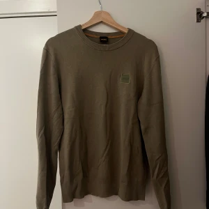 Hugo boss sweatshirt - Snygg olivgrön tröja från BOSS med rund halsringning och diskret logopatch på bröstet. Tröjan är i mjuk bomull och har långa ärmar samt ribbade muddar vid hals, ärmslut och nederkant. Perfekt för en clean och stilren look.