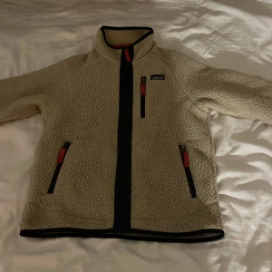 Beige fleecejacka från Patagonia L barnstorlek - Säljer en beige fleecejacka från Patagonia i barnstorlek L. Jackan har hel dragkedja, tre fickor med svarta och röda detaljer samt hög krage. Perfekt för kyliga dagar och riktigt skön att ha på sig.