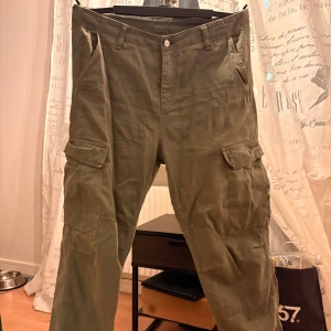 Olivgröna cargobyxor från Carhartt - Säljer ett par olivgröna cargobyxor från Carhartt med klassiska cargofickor på sidorna och bakfickor med lock. Byxorna har raka ben, bälteshällor och är tillverkade i slitstark bomull. Perfekta för en avslappnad streetwear-look.