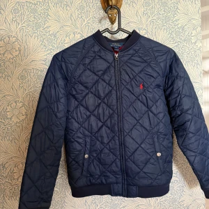 Marinblå quiltad bomberjacka Polo Ralph Lauren - Snygg marinblå quiltad bomberjacka från Polo Ralph Lauren med dragkedja framtill, två fickor med tryckknappar och klassisk röd logga på bröstet. Jackan har ribbade muddar och är helfodrad i rött. Perfekt för höst och vår.