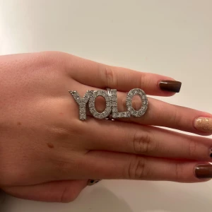 Glittrig YOLO-ring i silverfärg Ioaku  - Statementring med texten 'YOLO' täckt av glittrande stenar i silverfärg. perfekt för dig som vill sticka ut och visa att du lever livet fullt ut. Från Ioaku 