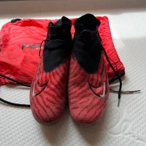 Nike Phantom GT2 Elite fotbollsskor röd/svart - Nike Phantom GT2 Elite fotbollsskor i rött och svart med coolt mönster och vita Swoosh-loggor. Skorna har hög strumpkrage, snörning och textilöverdel för grym passform och kontroll. Perfekta för dig som vill sticka ut på planen.
