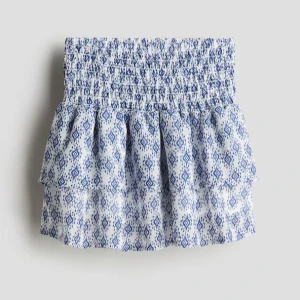 Blåvit mönstrad volangkjol - Supersöt kort kjol med blått mönster på vit botten. Kjolen har smock i midjan och dubbla volanger för extra volym. Tillverkad i tunn, luftig bomull med innerfoder. Perfekt för varma dagar när du vill ha en chill och fräsch look.  Storlek 158 (barn) men den passar jätte bra på mig som är storlek XS\XSS