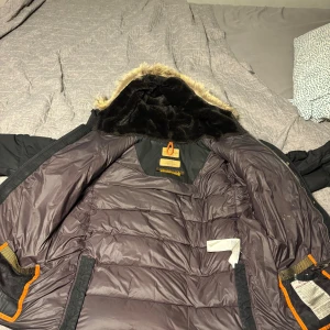 Svart pufferjack från Parajumpers - Säljer en svart pufferjacka från Parajumpers med huva och fuskpälsdetalj. Jackan har flera fickor, patch på ärmen och orangea detaljer på insidan. Perfekt för kalla dagar med sin tjocka vaddering och snygga design. Den har något på insidan vet ej vad det är men man tänker inte så mycket på det.