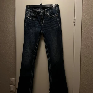 Blå bootcut jeans från Miss Me, stl 27 - Snygga mörkblå jeans från Miss Me med tydliga kontrastsömmar och dekorerade bakfickor med knappar och broderade detaljer. Modellen har bootcut-ben och klassisk femficksdesign. Använd mycket sparsamt. Den ligger också ute på Vinted! Tveka inte att fråga mig vid minsta lilla fundering❣️