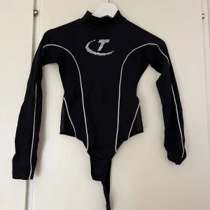 Svart långärmad topp från Trapstar - Svart långärmad topp/body från Trapstar med hög hals och vita kontrastlinjer samt mest på sidorna. Framsidan har ett glittrigt T i strass. Sitter som en XS