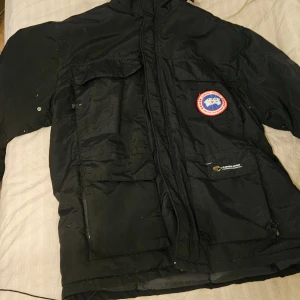 Svart dunjacka från Canada Goose - Svart dunjacka från Canada Goose med klassisk logga på bröstet och broderad etikett på ryggen. Jackan har stora fickor framtill, huva och dragkedja. Yttermaterialet är i slitstark polyester och fodret är fyllt med dun för extra värme. Perfekt för kalla vinterdagar.
