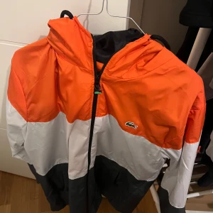 Färgstark vindjacka från Lacoste - Säljer en snygg vindjacka från Lacoste i orange, vitt och svart. Jackan har huva, dragkedja framtill och klassisk Lacoste-logga på bröstet. Perfekt för dig som vill sticka ut med en sportig och fräsch look.