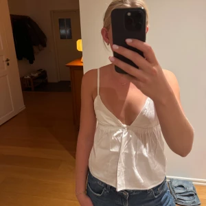 Vit topp med smala axelband - Säljer en vit topp med smala axelband och djup v-ringning. Toppen har en luftig passform och är tillverkad i bomull, perfekt för varma dagar. Enkel och stilren design som är lätt att matcha med jeans eller kjol. Man ser inte igenom, kan därför användas utan bh. 