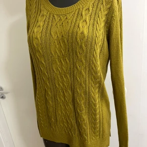 Olivgrön stickad tröja från Lindex - Snygg olivgrön stickad tröja från Lindex med flätstickat mönster framtill. Tröjan har rund halsringning, långa ärmar och ribbade muddar. Perfekt för lager-på-lager och chill höststil.