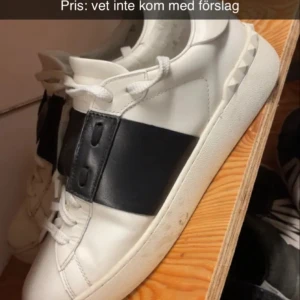 Valentino Garavani Open sneakers vit/svart - Säljer ett par snygga Valentino Garavani Open sneakers i vitt skinn med bred svart läderdetalj över vristen. Klassisk rund tå, vita snören och ikoniska nitar längs sulan. Perfekta för dig som vill ha en clean men ändå lyxig look.