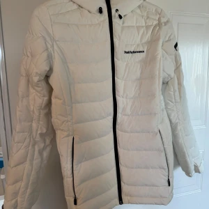 Vit pufferjacka från Peak Performance - Säljer en snygg vit pufferjacka från Peak Performance med huva och svarta detaljer. Jackan har dragkedja framtill, två fickor med dragkedja och diskret logga på bröstet. Perfekt för kalla dagar när du vill hålla stilen.