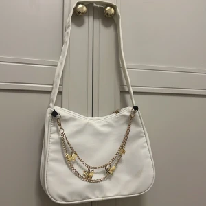 Vit axelväska med guldkedja och fjärilar - Snygg vit axelväska i syntetmaterial med mjuk form och dragkedja upptill. Framsidan är dekorerad med en guldfärgad kedja och små fjärilar som ger väskan en unik och trendig look. Perfekt storlek för det viktigaste och enkel att bära över axeln.