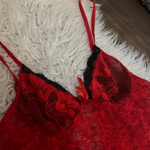 Röd spetsig nattlinne med svarta detaljer - Säljer en röd spetsig nattlinne med svarta spetsdetaljer vid bysten och små rosetter framtill. material är genomskinlig spets med blommigt mönster och smala justerbara axelband. Knappt använd! 
