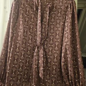 Brun mönstrad blus Pernilla Wahlgren - Snygg brun blus från Pernilla Wahlgren med unikt grafiskt mönster i beige. Blusen har lång ärm, knytband vid halsen och är gjord i ett mjukt, glansigt material som känns lyxigt. Perfekt för dig som gillar att sticka ut med coola detaljer och mönster.
