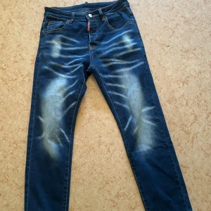 Blå Dsquared2 jeans med slitningar - Säljer ett par blå jeans från Dsquared2 i storlek 44. Jeansen har snygga slitningar och ljusa tvättade partier framtill, klassisk femficksmodell och Dsquared2-logga på röd etikett vid fickan. Materialet är jeans med kontrastsömmar och smal passform. Helt nya endast använt 2 gånger taggar finns kvar  