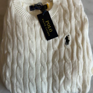 Vit kabelstickad tröja Polo Ralph Lauren - Kabelstickad vit tröja från Polo Ralph Lauren med klassisk rund halsringning och korta ärmar. Tröjan har broderad logga på bröstet och ribbade muddar. Perfekt för en clean och stilren look.
