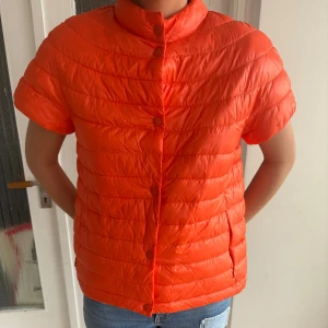 Orange pufferjacka från Italien  - Säljer en kortärmad orange pufferjacka från Joseline Firenze. Jackan är quiltad, har hög krage och knäpps med knappar framtill. Tillverkad i 100% polyester och är lätt och smidig, perfekt för lager-på-lager. Made in Italy. Köpt på en liten butik i Italien! Aldrig använt. 