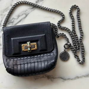 Svart axelväska från Lanvin  - Snygg svart axelväska från Lanvin i skinn med cool kedjerem i metall. Väskan har ett unikt flätat mönster på framsidan och ett dekorativt spänne i guld och silver. Perfekt storlek för det viktigaste och med en exklusiv känsla.