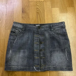 Grå jeanskjol med knappar från ICHI - Snygg grå jeanskjol från ICHI med slitna detaljer och knappar framtill. Kjolen har klassiska fickor både fram och bak samt bälteshällor. Perfekt för dig som gillar en cool och avslappnad stil.