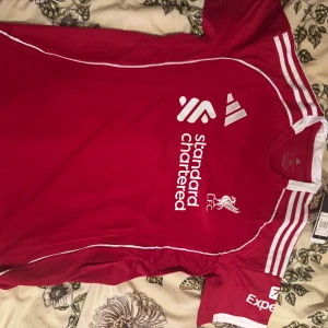 Liverpool FC röd matchtröja Adidas - Liverpool FCs officiella matchtröja i rött från Adidas med vita detaljer och tryck. Tröjan har korta ärmar, rund hals och klubbmärke samt sponsorloggor på bröstet. På ryggen står det ISAK och nummer 9 i vitt. Tillverkad i lätt och ventilerande material.