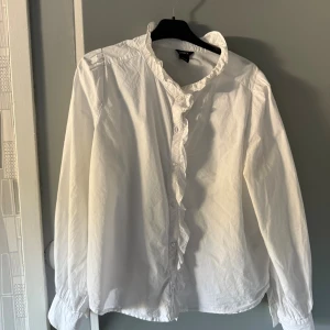 Vit volangskjorta från Lindex - Vit skjorta från Lindex med volangkant längs kragen och knäppningen framtill. Skjortan har långa ärmar och är tillverkad i mjuk bomull. Perfekt för dig som gillar en romantisk och stilren look.