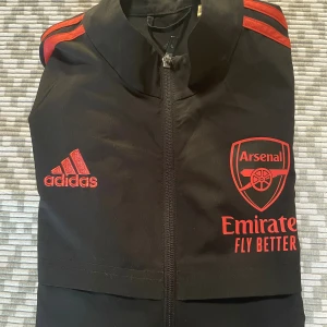 Arsenal x Adidas svart vindjacka S - Svart vindjacka från Adidas i samarbete med Arsenal, med röda detaljer och loggor på bröstet. Jackan har dragkedja framtill, långa ärmar och röda ränder på axlarna. Perfekt för dig som älskar fotboll och vill ha en sportig look. Jackan är knappt använd