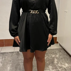 Svart kort satinklänning med bälte - Stilren svart kort klänning i satinliknande material med långa ärmar och markerad midja. Klänningen har ett snyggt bälte med kedjedetalj framtill och knytning bak. Perfekt för dig som gillar en elegant och trendig look.