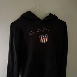 Svart hoodie från GANT med tryck - Svart hoodie från GANT med klassiskt tryck och sköld på bröstet. Tröjan har huva och långa ärmar, perfekt för en chill och avslappnad stil. Mjuk och bekväm med känguruficka framtill.