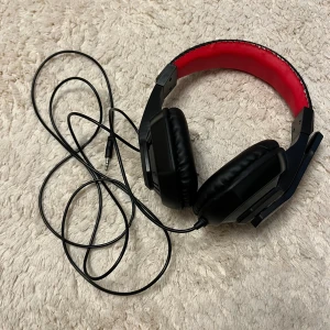 Kabelanslutet spelheadset med mikrofon - Bekvämt over-ear spelheadset med kabel och inbyggd mikrofon. Svart design med röd vadderad bygel. 3,5 mm kontakt. Synligt slitage på ena kåpan, men i övrigt gott skick. Perfekt för gaming eller videosamtal.
