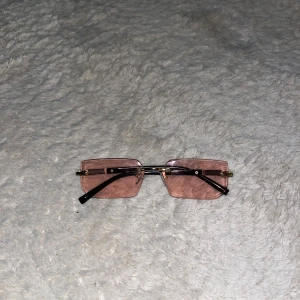 Rektangulära solglasögon med rosa glas - Snygga rektangulära solglasögon med tunna svarta skalmar och guldiga detaljer. Glasen är ljusrosa och ger en trendig vibe. Perfekta för dig som vill sticka ut med en unik accessoar. Lätta och stilrena, passar till många olika looks.