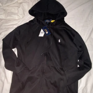 Svart hoodie från Polo Ralph Lauren - Svart hoodie från Polo Ralph Lauren med dragkedja framtill och klassisk huva. Tröjan har snörning vid huvan och den ikoniska vita loggan broderad på bröstet. Perfekt för en chill och stilren look.