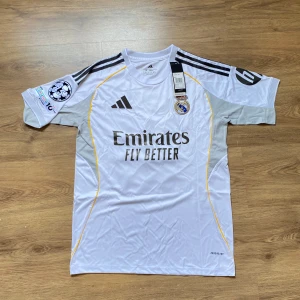 Real Madrid tröja - Real Madrid fotbollströja med Mbappé och nummer 10 på ryggen. Vit med svarta detaljer, gula linjer och grå partier på sidorna. Broderat klubbmärke, Champions League-märke och UEFA Foundation-patch. Tillverkad i lätt och ventilerande polyester (AEROREADY).