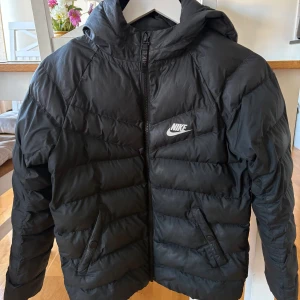 Svart pufferjacka från Nike med huva - Säljer en svart pufferjacka från Nike med huva och dragkedja framtill. Jackan har två snedställda fickor med knappar och klassisk Nike-logga på bröstet. Säljer för 400 kr, finns i Kalmar. Kan skickas mot fraktkostnad. 