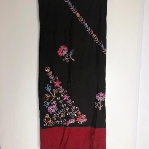 Svart och vinröd halsduk med blommor - En lång svart halsduk med vinröd kant och broderade blommor i flera färger. Halsduken har fransar i ändarna och ett färgglatt, broderat blommönster som ger en unik och kreativ vibe. Perfekt accessoar för att lyfta din outfit.