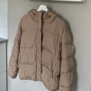 Beige pufferjacka med huva - Säljer en beige pufferjacka med oversized passform och huva. Jackan har dragkedja och knappar framtill samt snörning i huvan. Perfekt för kalla dagar och riktigt skön att bära. Fodrad och quiltad för extra värme.
