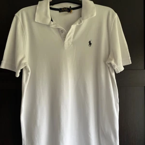 Vit pikétröja från Polo Ralph Lauren - Vit klassisk pikétröja från Polo Ralph Lauren med korta ärmar och broderad logga på bröstet. Tröjan har krage och knappar framtill, tillverkad i mjuk bomull. Perfekt för en clean och stilren look.