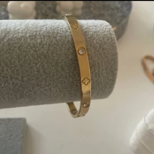 Guldigt armband med stenar - Snyggt armband i guldig metall med infällda klara stenar och ingraverade fyrklöverliknande symboler runt om. Stilren och modern design som ger en lyxig känsla till din outfit.