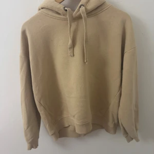 Beige hoodie med huva och snörning - En stilren beige hoodie med huva och snörning. Tröjan har en avslappnad passform, ribbade muddar vid ärmslut och nederkant samt lång ärm. Perfekt för en chill och enkel look. Aldrig använt den